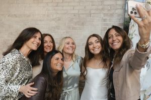 A group of friends laugh and smile together while taking selfies against a white brick wall.
