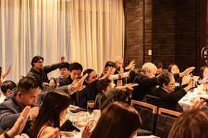 A large group of people in a conference room raise their hands during an interactive presentation session.