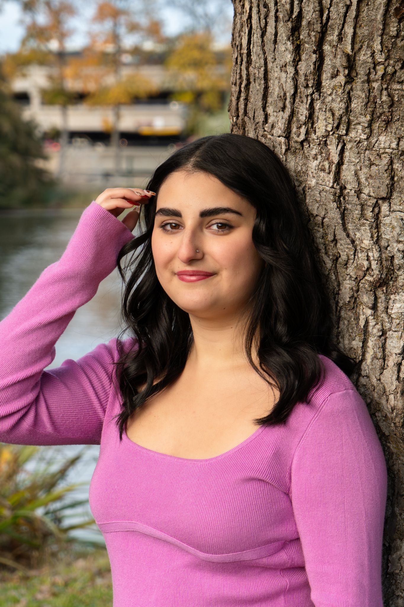 A person in a pink sweater poses against a tree trunk in an autumn outdoor setting with natural lighting.
