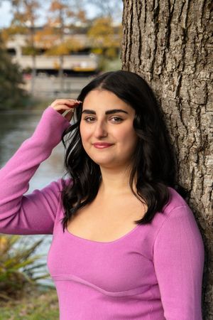 A person in a pink sweater poses against a tree trunk in an autumn outdoor setting with natural lighting.