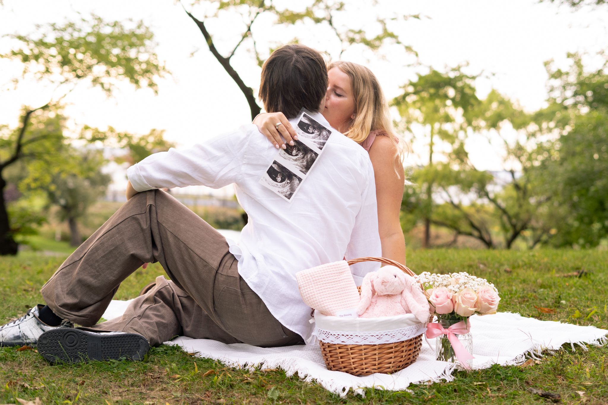 Couple sitting on blanket in park sharing ultrasound photos with pink baby items in wicker basket, pregnancy announcement.