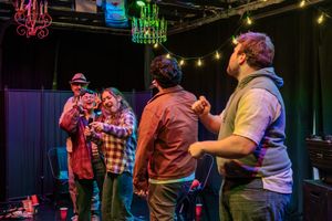 A group of performers interact on a dark stage illuminated by colorful theatrical lighting.