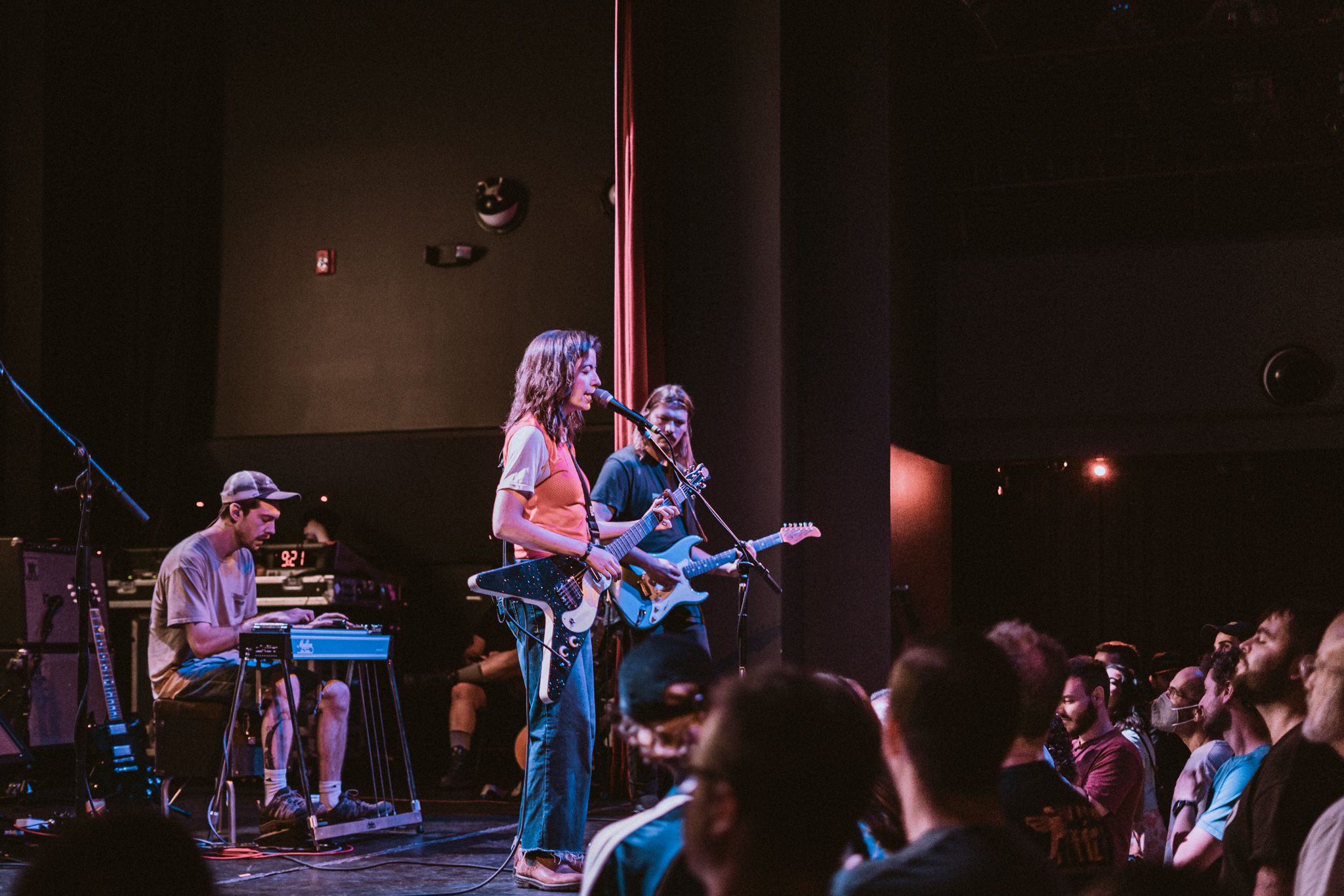 Live band performing on stage with electric guitar and keyboard, engaging a seated audience in a dimly lit venue.