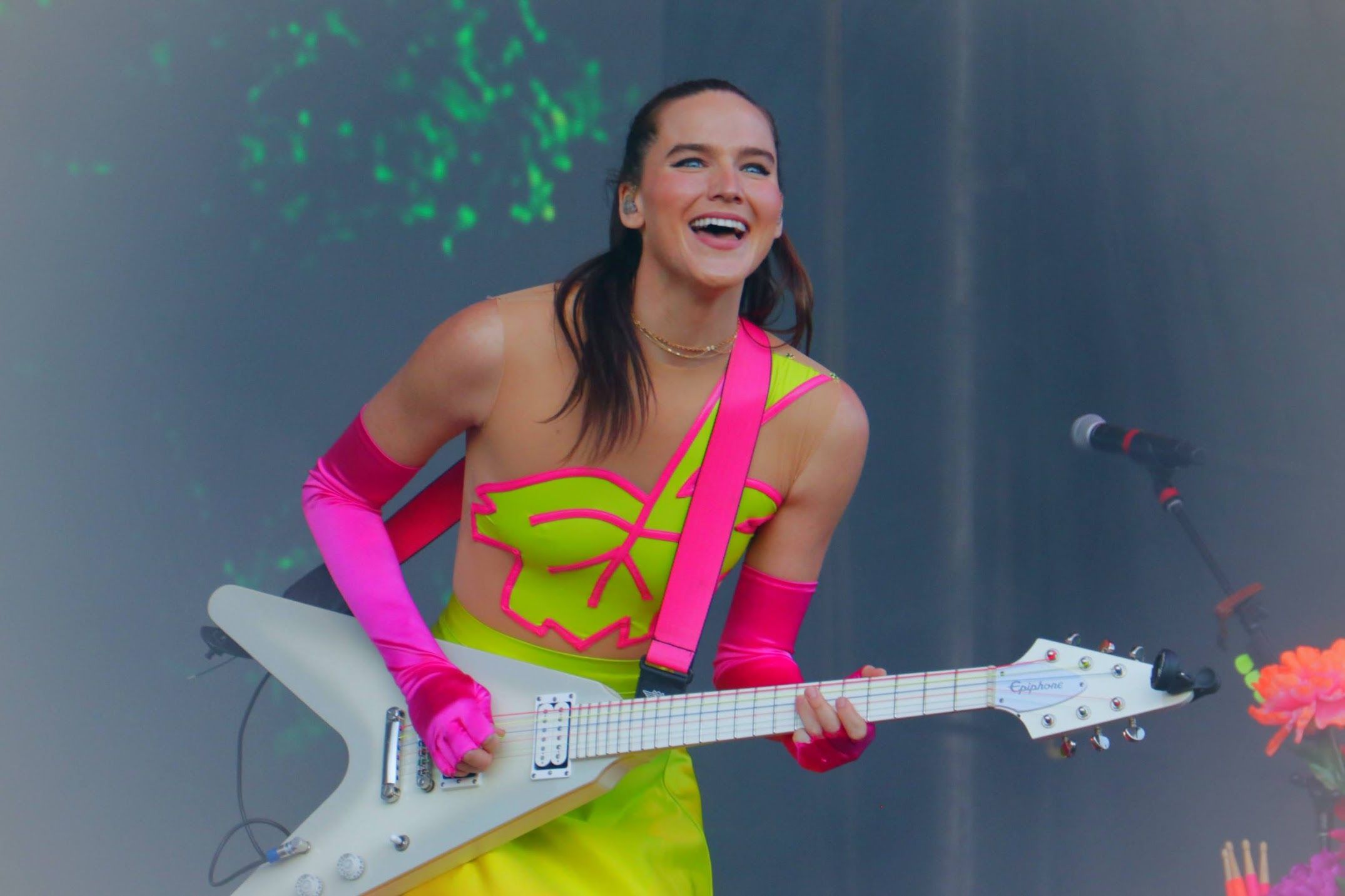 Performer in bright neon yellow dress and pink gloves playing electric guitar on stage with green lighting.