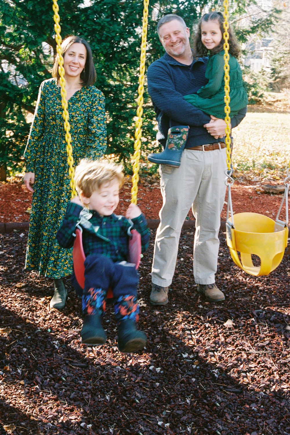 Family poses by the swing set in their backyard in the sunshine in the fall in Portland, Maine.