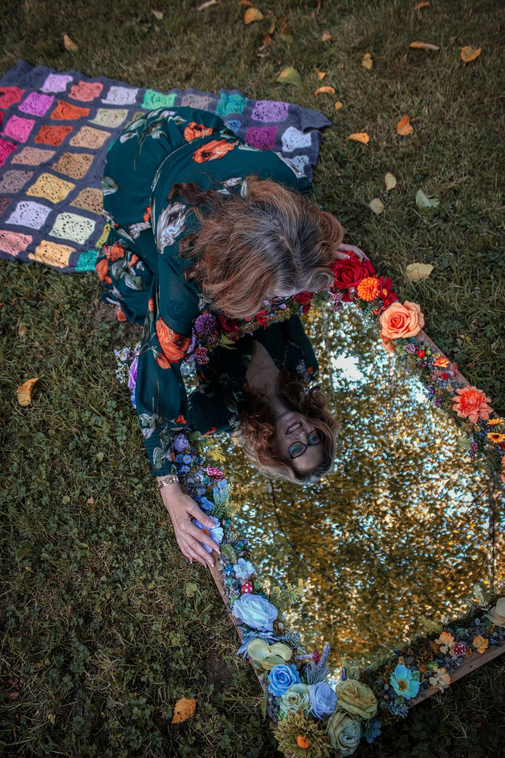 A person lies on grass on a blanket with a decorated mirror.