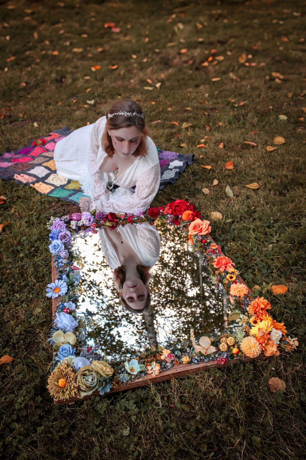 A dreamy outdoor scene with someone in white lying on a mirror surrounded by colorful flowers on autumn grass.