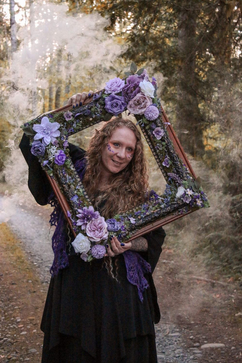 Someone holding a vintage frame decorated with purple flowers in a forest setting.