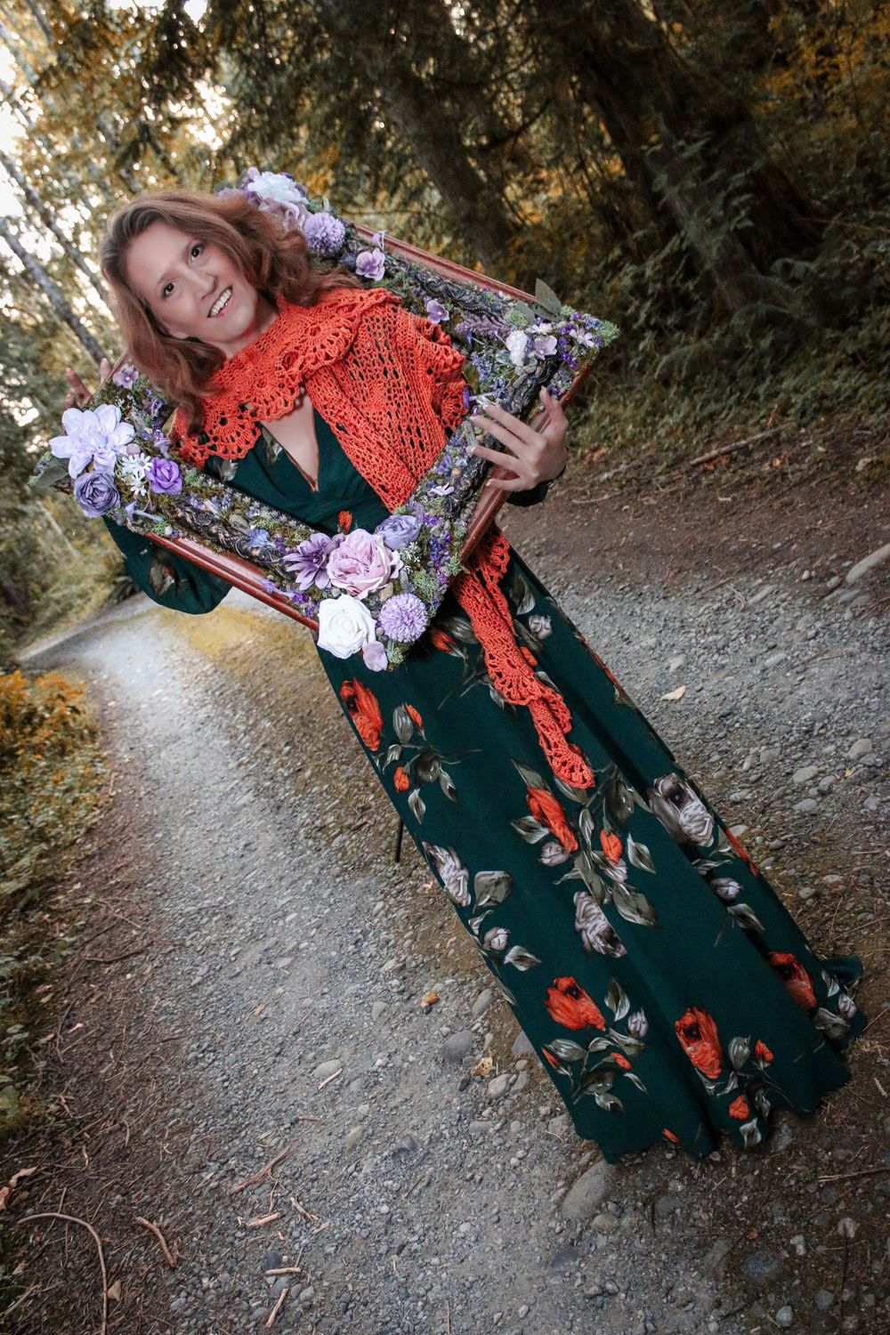 A flowing floral dress is paired with a bright orange lace shawl while holding a decorated mirror in a forest setting.