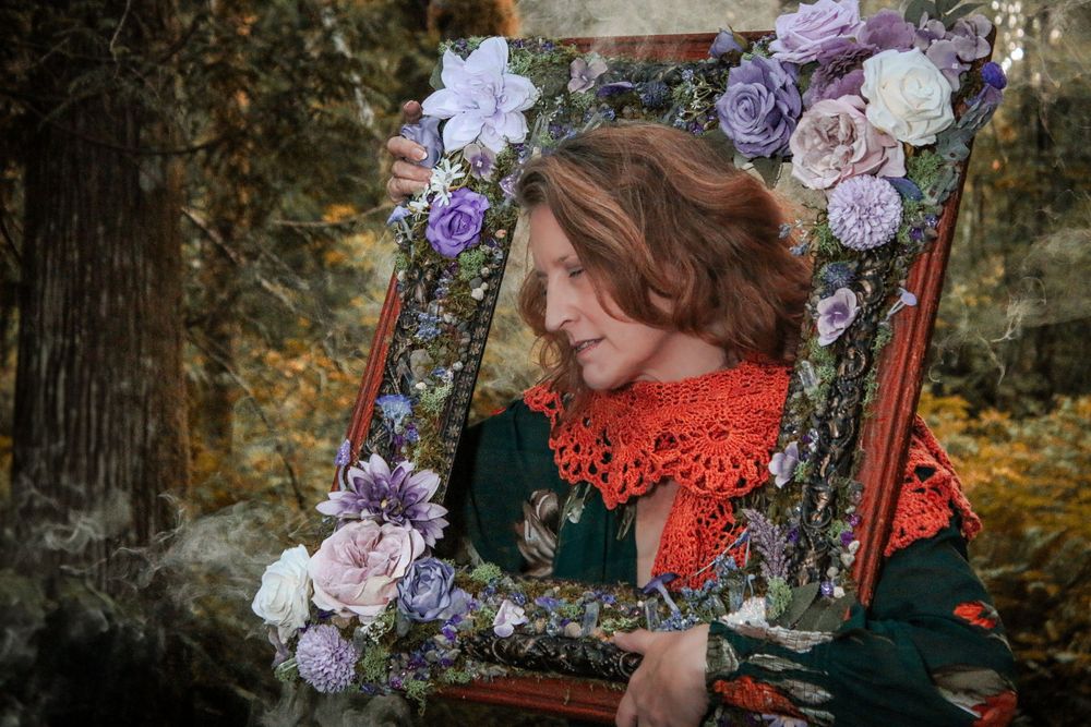 A decorative vintage frame adorned with purple and pink flowers sits in a forest setting during autumn.