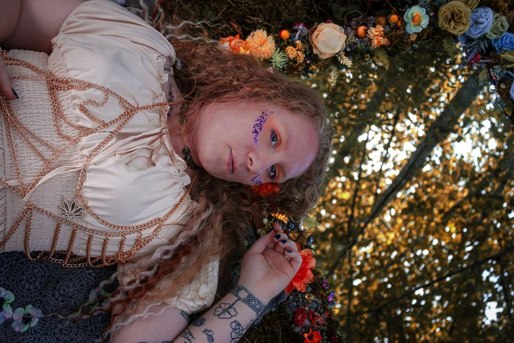 A person in a vintage-style dress poses on a decorated-with-flowers mirror, which reflects the canopy of trees above.