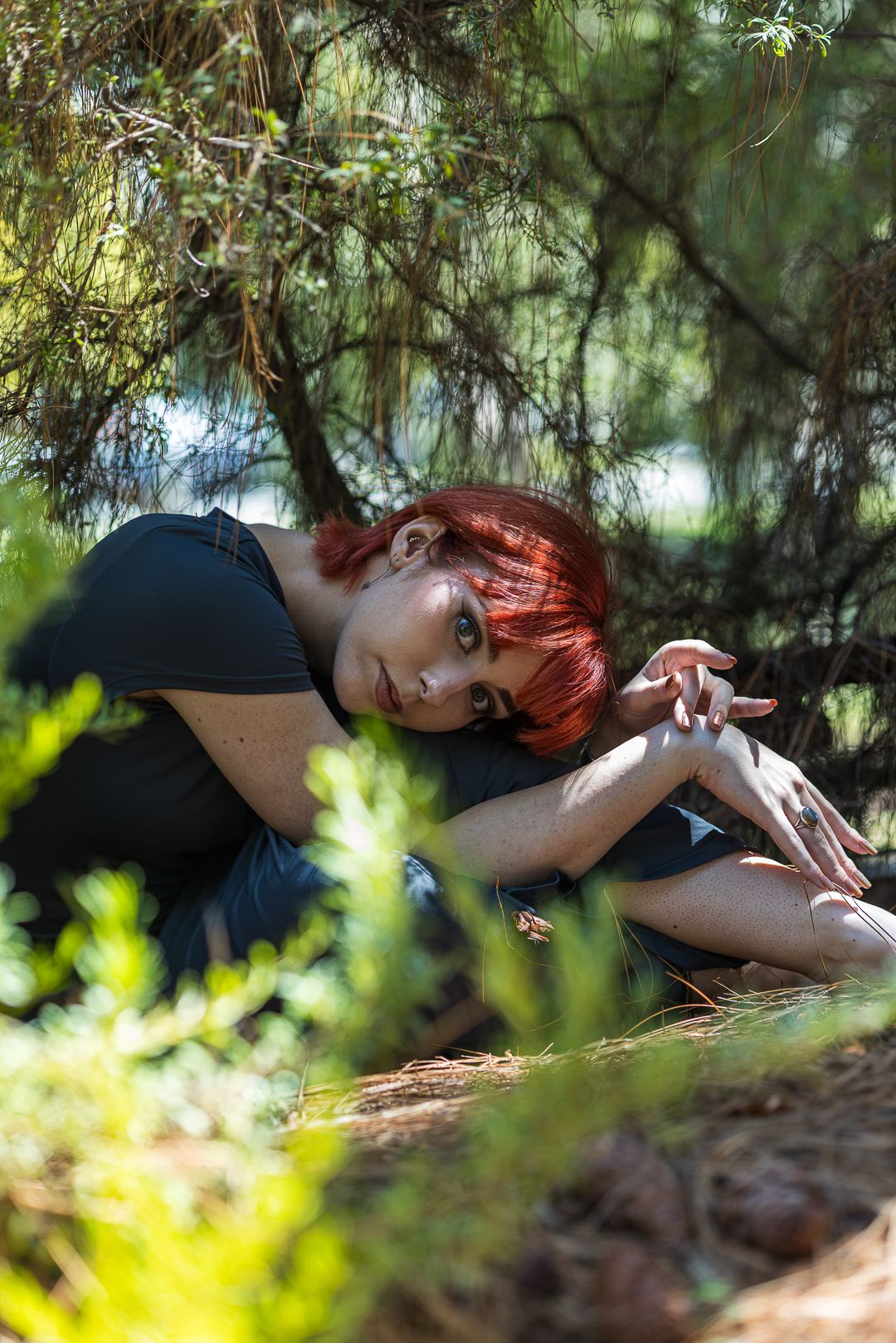 A model with striking red hair relaxes in the woods, featured in a model photoshoot surrounded by nature.