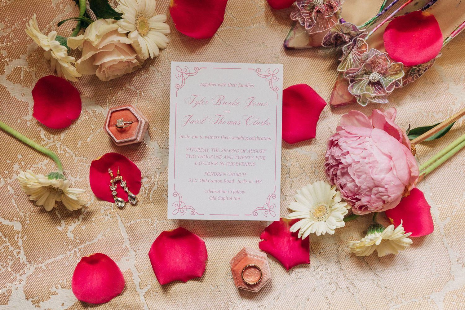 Wedding invitation surrounded by pink peonies, red rose petals, and gold rings on a blush colored surface.