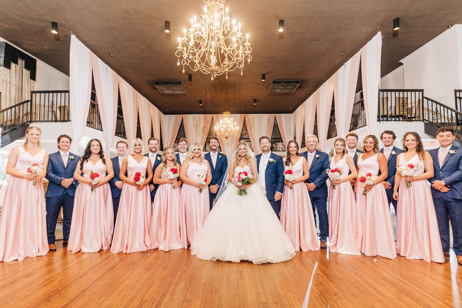 Wedding party in navy and blush pink attire pose in elegant ballroom with crystal chandelier overhead.