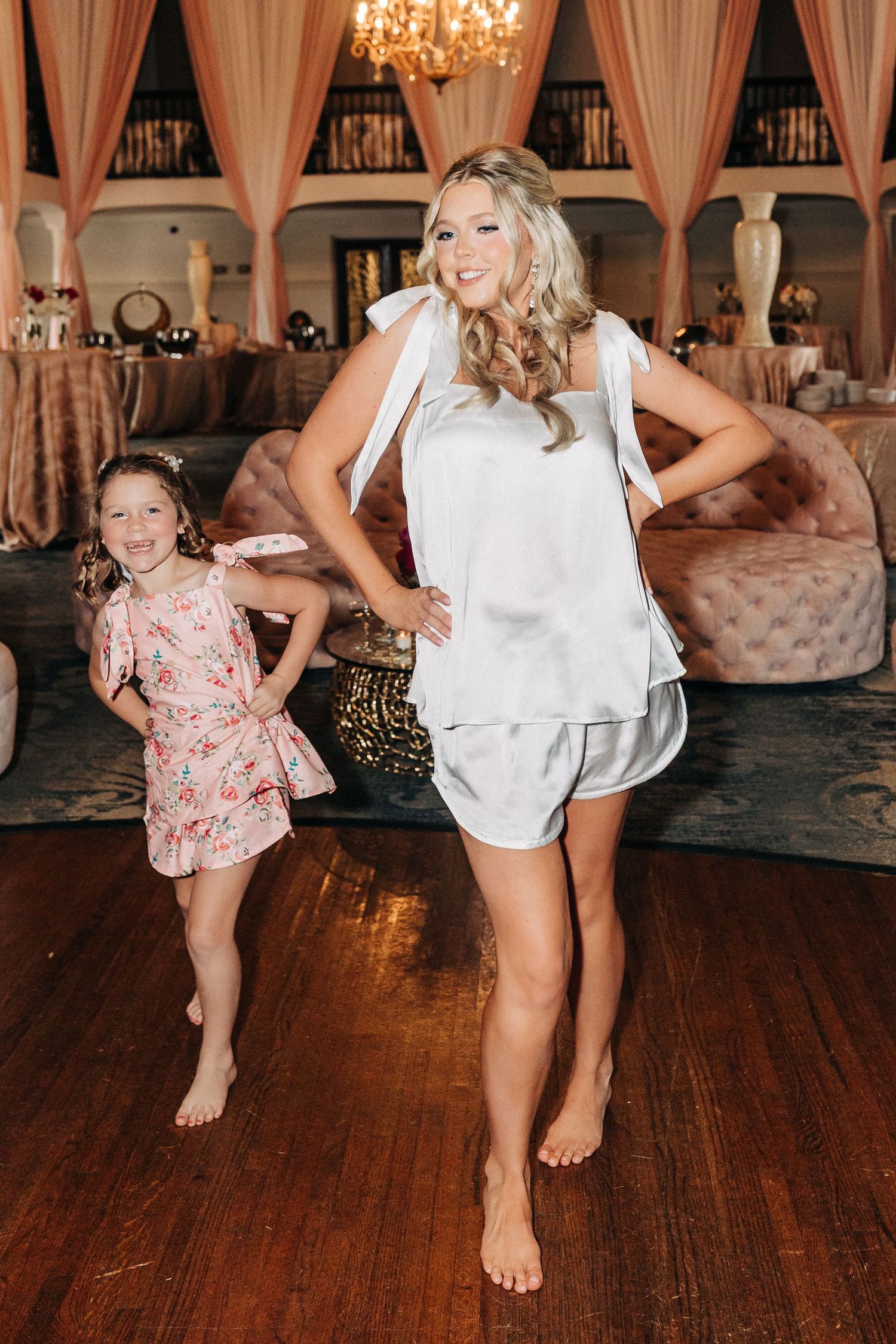 Two people dancing barefoot on wooden floor in elegant venue with chandelier and draped fabric decor.