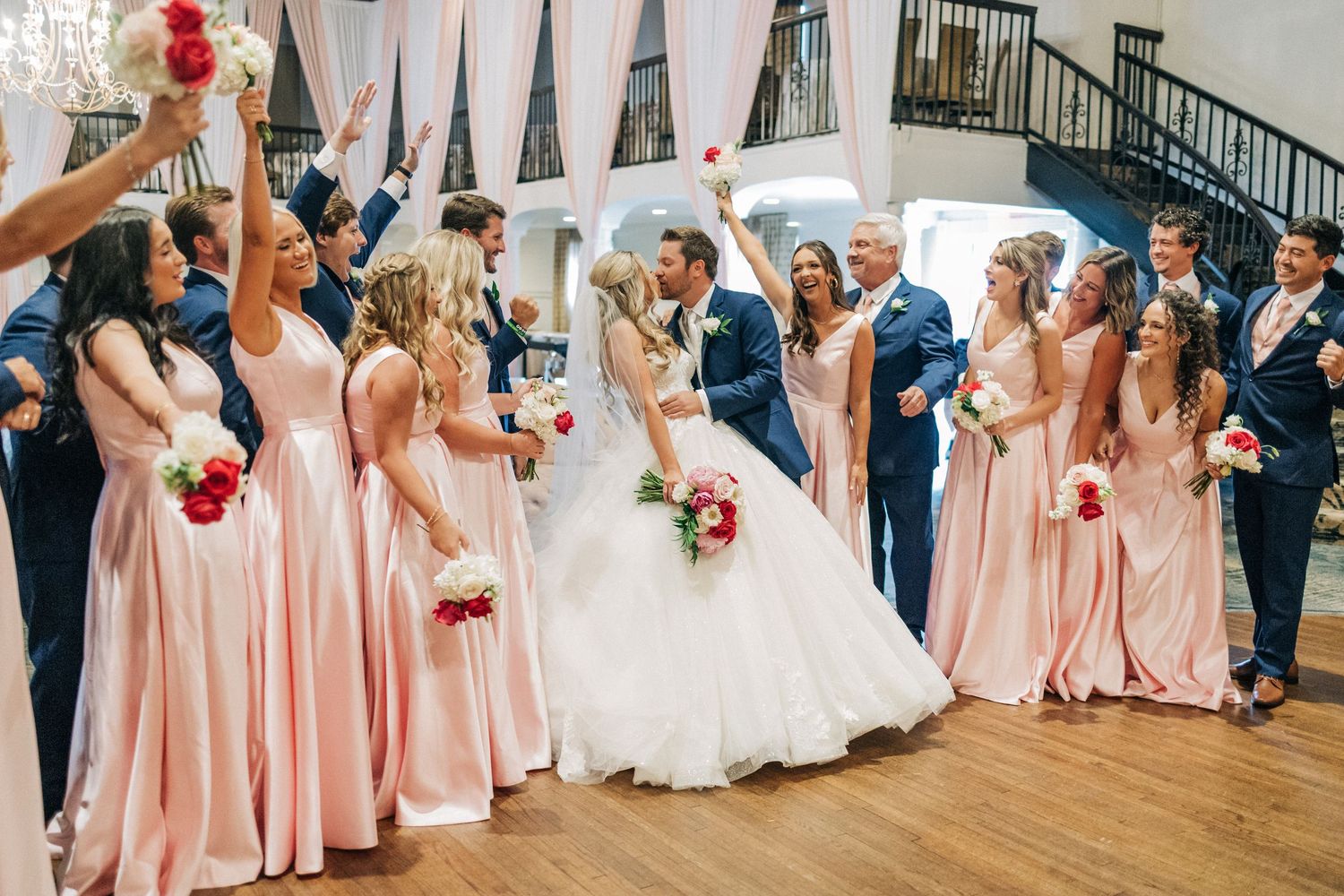 Wedding party celebrates with bridesmaids in pink dresses and groomsmen in navy suits on wooden dance floor.