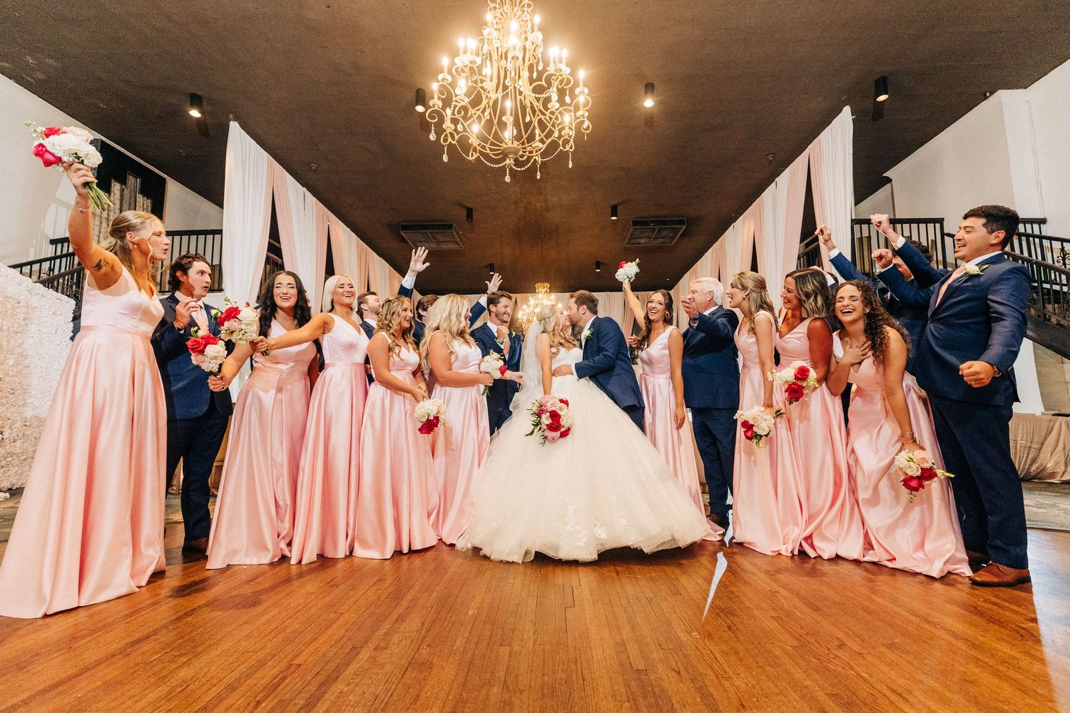 Wedding celebration under crystal chandelier with bridal party in blush pink gowns and navy suits.