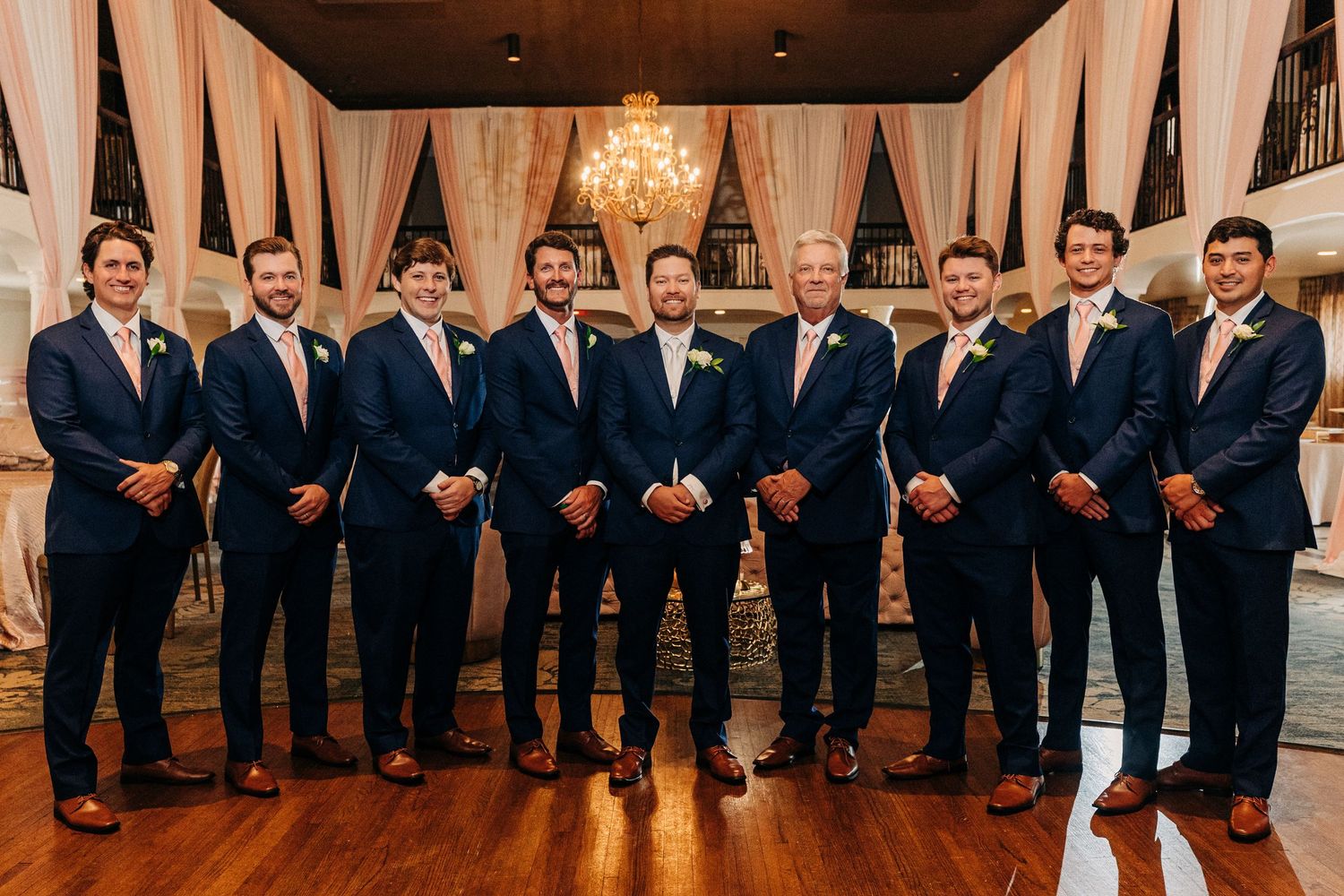 A group of groomsmen in matching navy suits stand together in an elegant venue with white drapery and chandeliers.