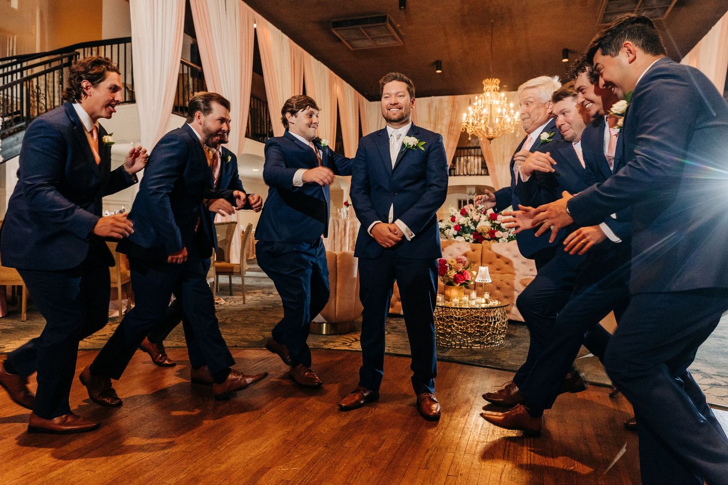 Groomsmen in navy suits dance energetically on a wooden dance floor at an elegant wedding reception venue.