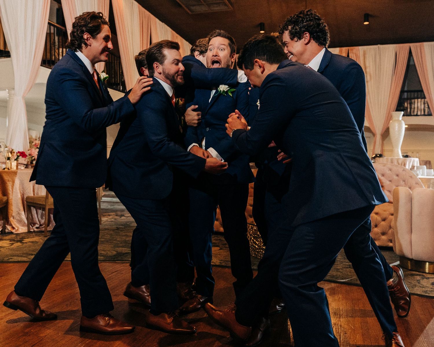A group of groomsmen in navy suits share an energetic celebration moment on the dance floor at a wedding reception.