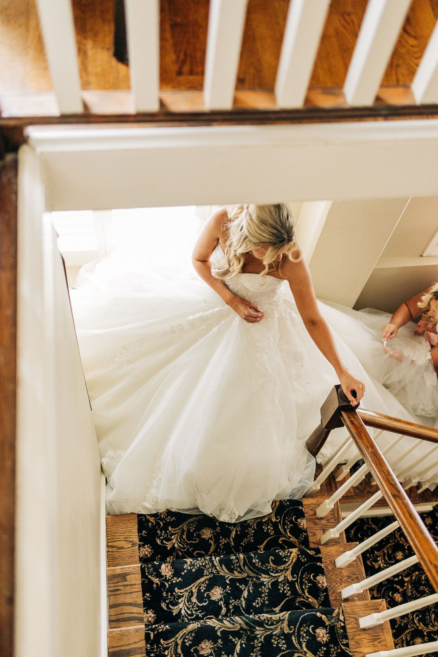 A bride in a flowing white wedding gown descends an elegant staircase with an ornate patterned carpet.