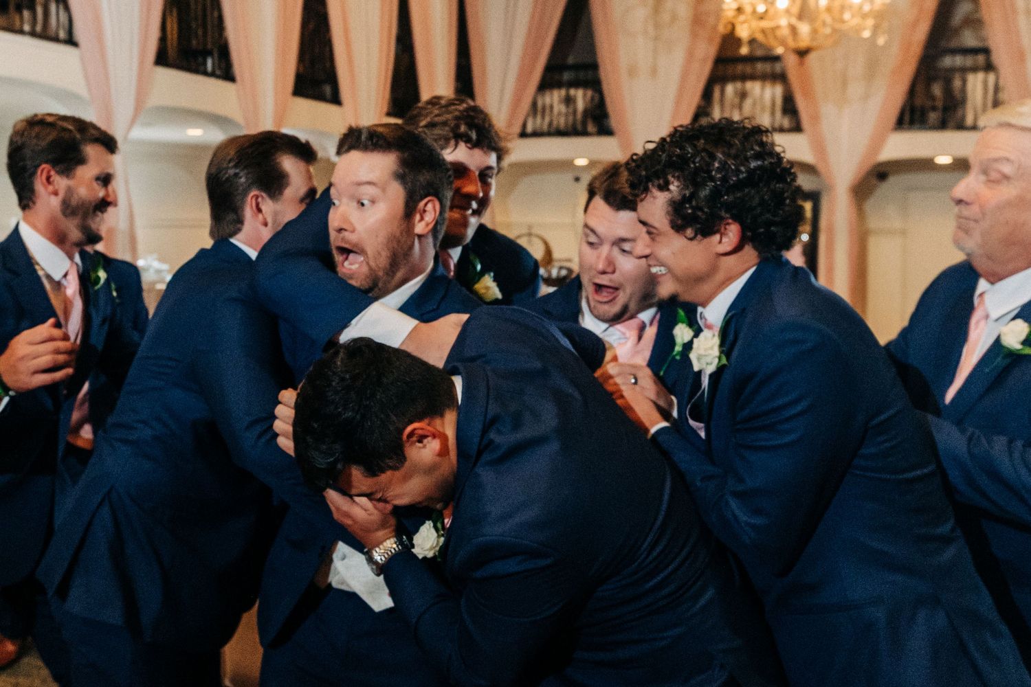 A group of groomsmen in navy blue suits share a candid laughing moment together at a formal wedding reception.