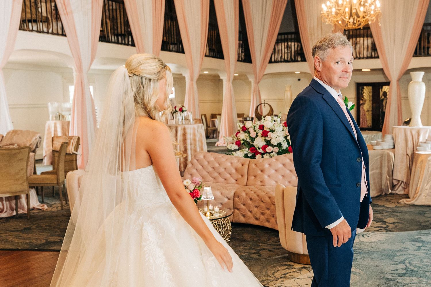 A romantic wedding first look moment takes place in an elegant ballroom with crystal chandeliers and white decor.