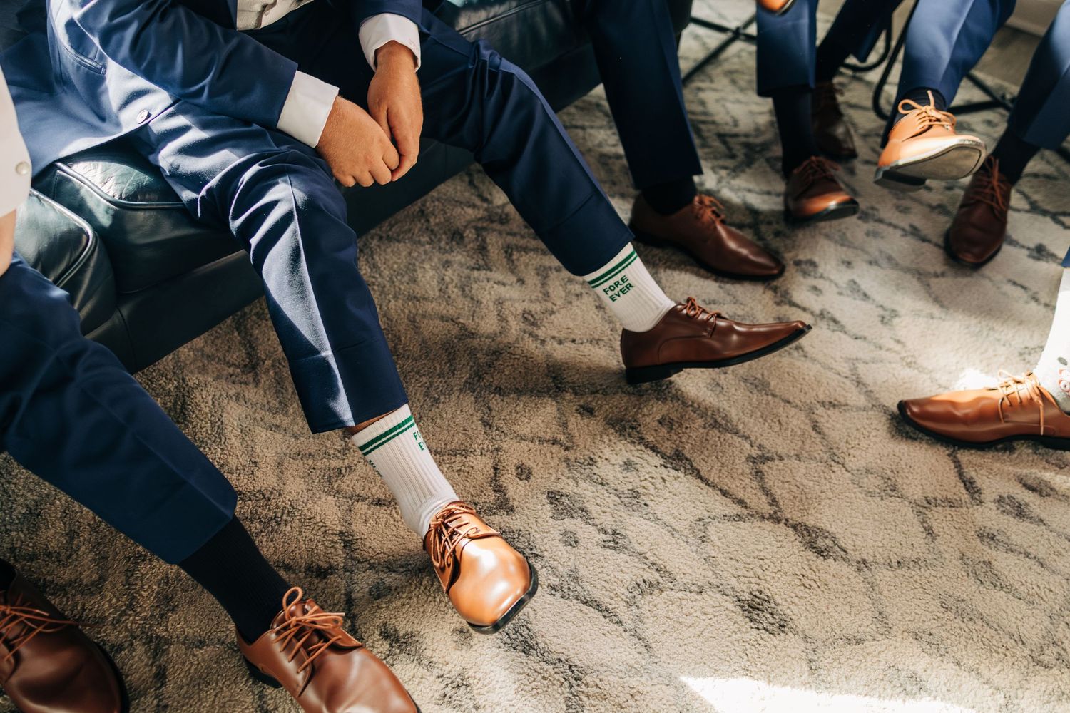 Row of wedding party groomsmen showing off colorful dress socks and brown leather shoes on cobblestone ground.
