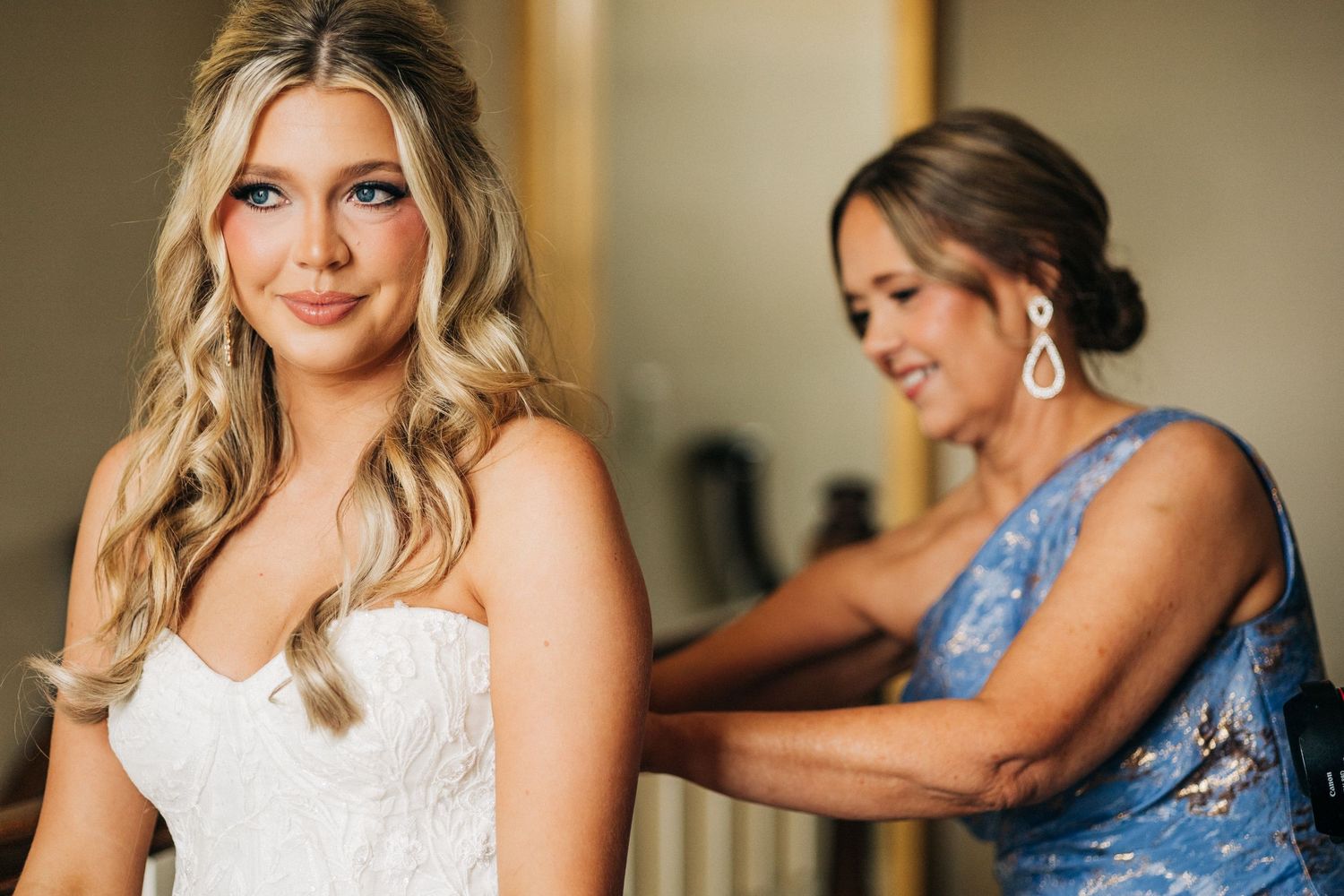A tender moment between someone in a white lace wedding dress and a person in blue helping with final adjustments.