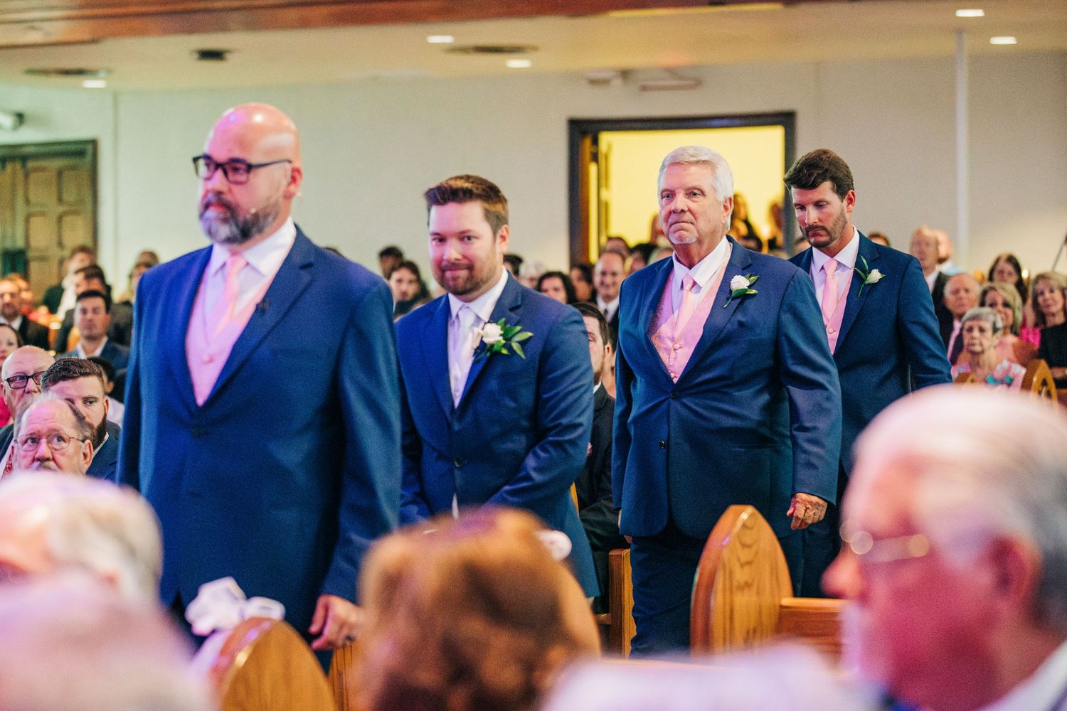 Wedding attendees wearing matching pink ties and navy suits stand in a ceremony space with a full audience.