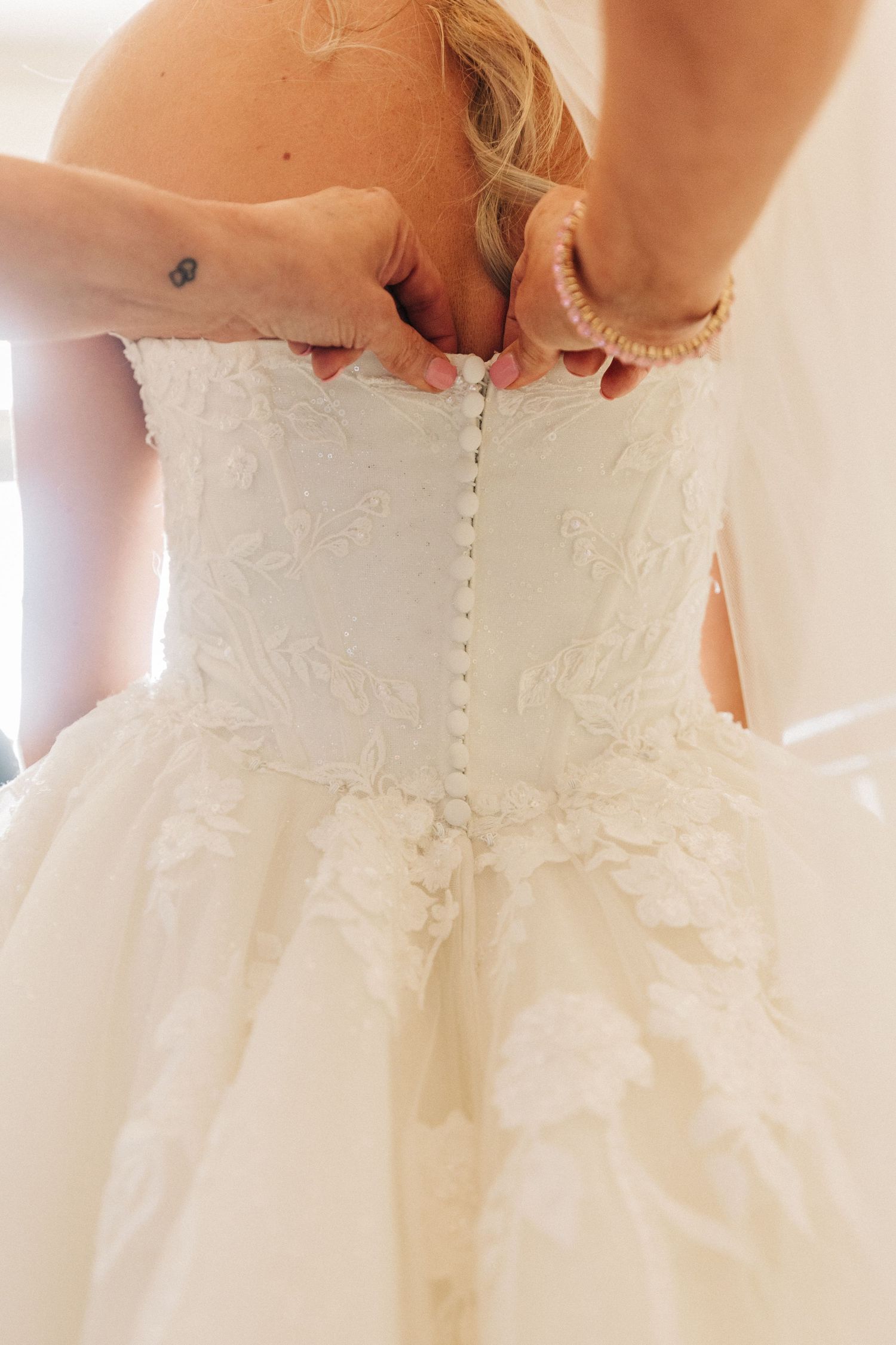 Close-up view of someone fastening the back of a white lace wedding dress with delicate buttons.
