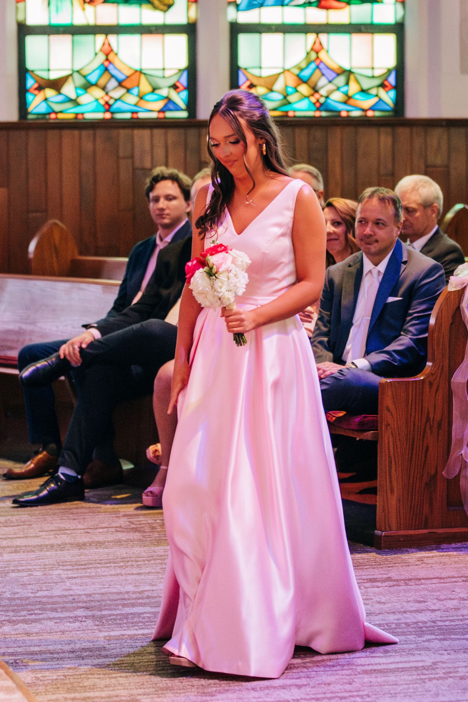 A bridesmaid in a flowing pink gown walks down the church aisle with stained glass windows visible in the background.