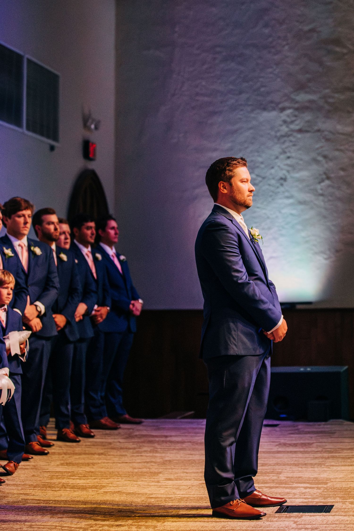 A groom stands at attention during a wedding ceremony while groomsmen line up against a white wall.