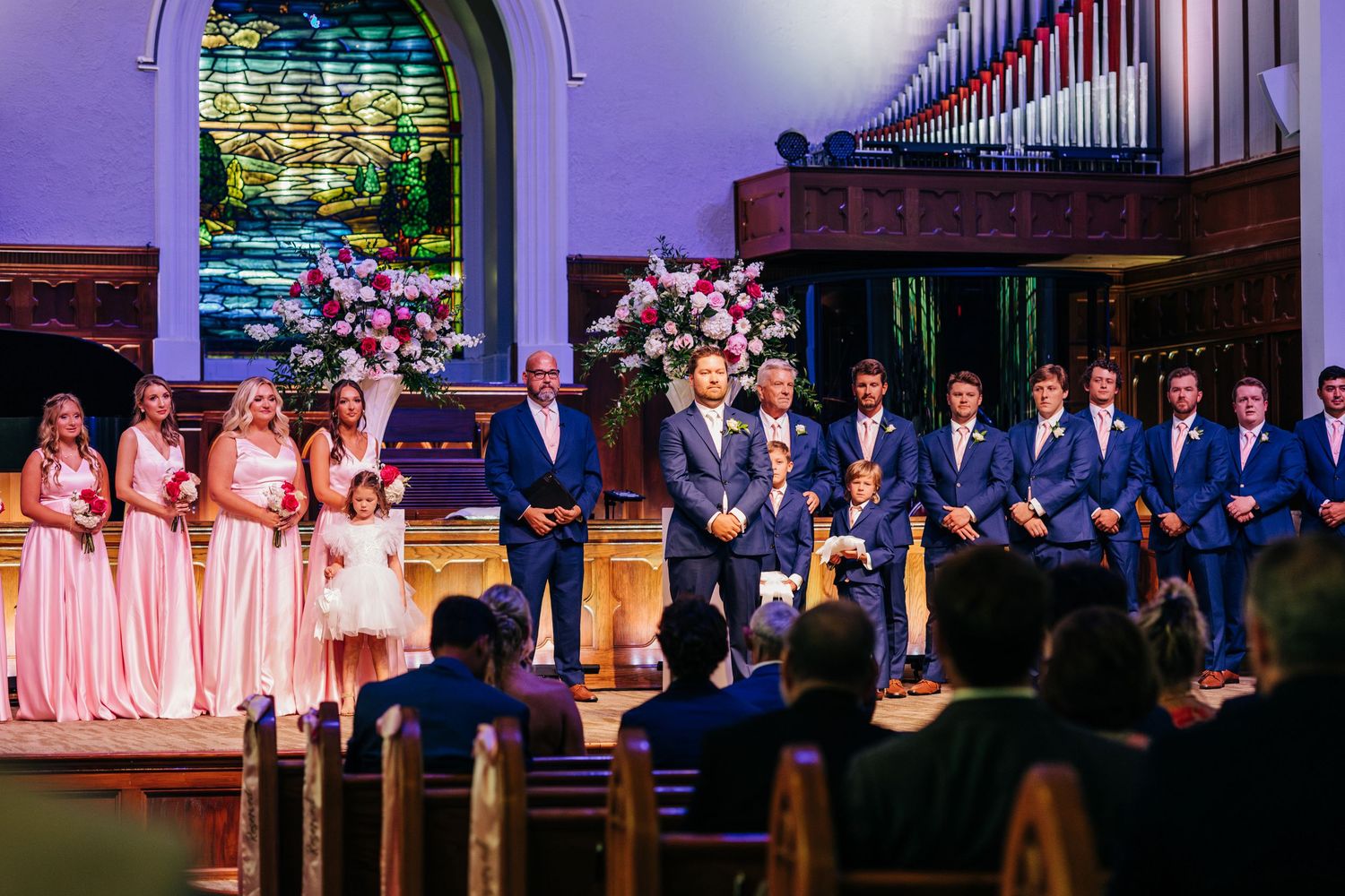 A wedding party lineup stands in a row at an elegant venue under purple and pink lighting.