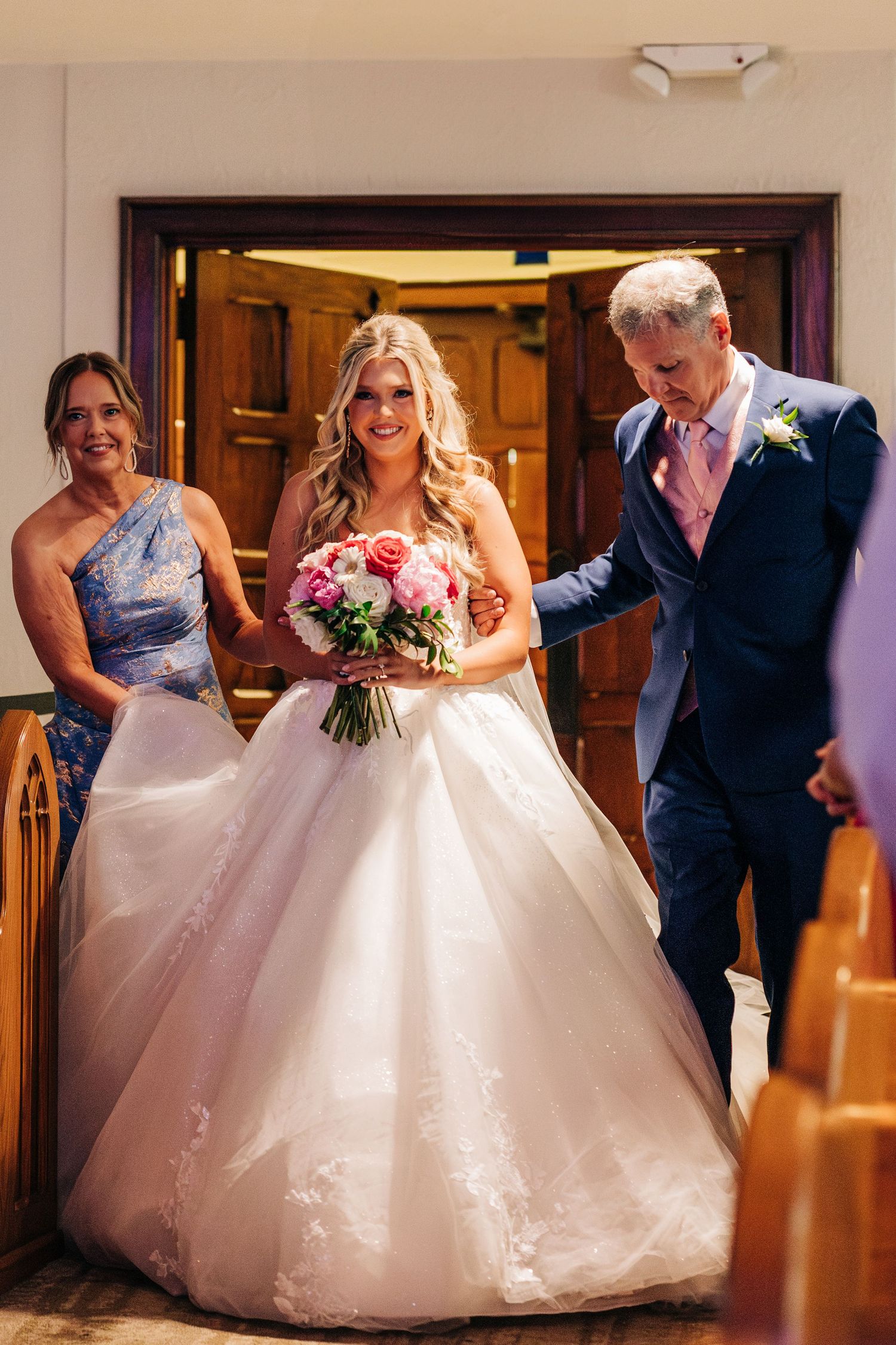 Wedding party entering through wooden doors while bride holds beautiful pink floral bouquet and wears a white ballgown.