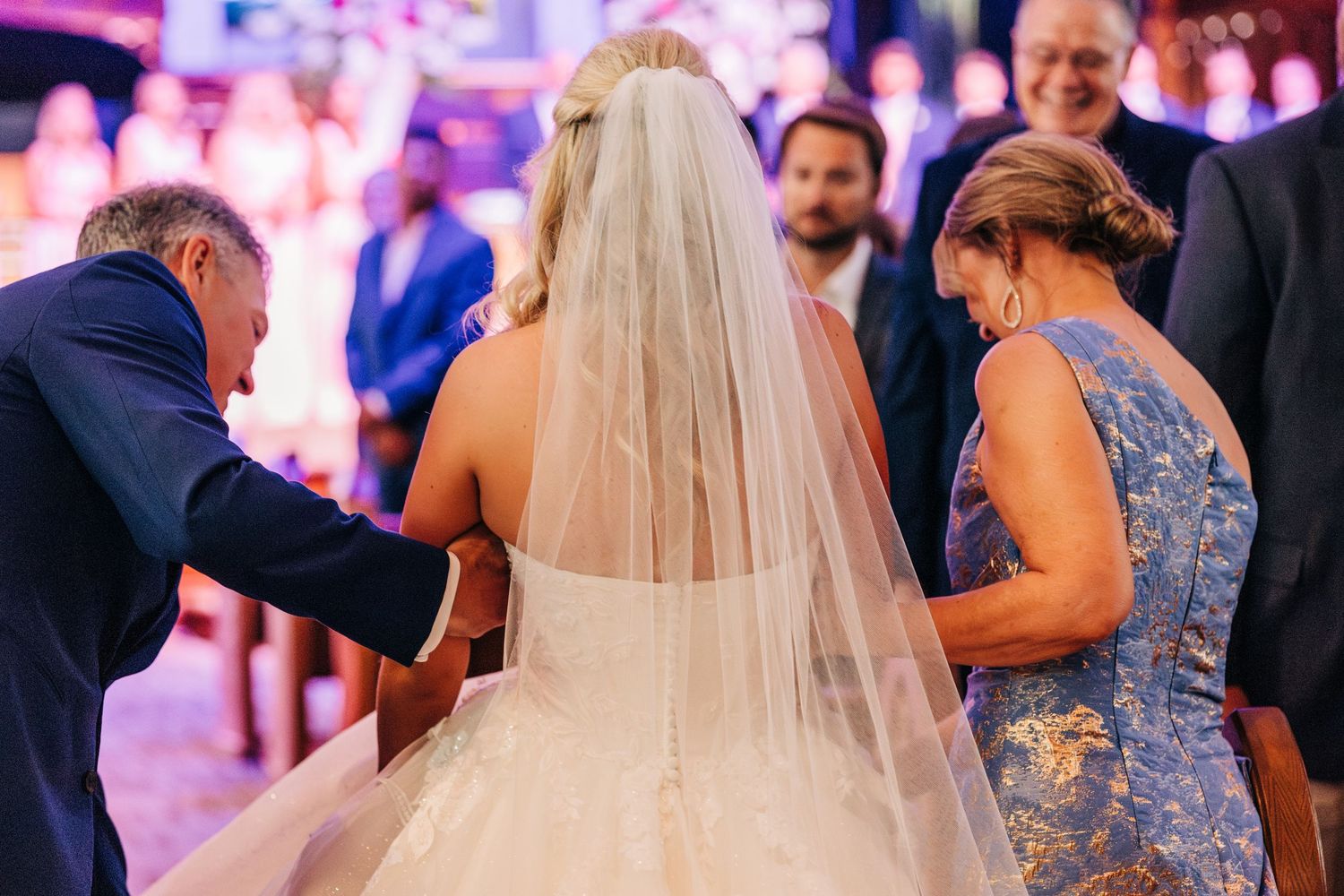 Wedding guests help adjust a bride's flowing veil during an evening reception with purple lighting.