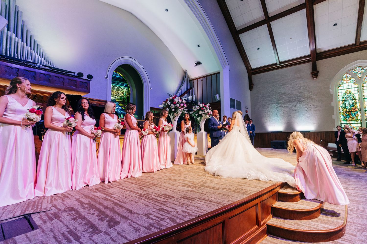 Wedding ceremony in a church with bridesmaids in pink dresses standing on a raised platform near the altar.