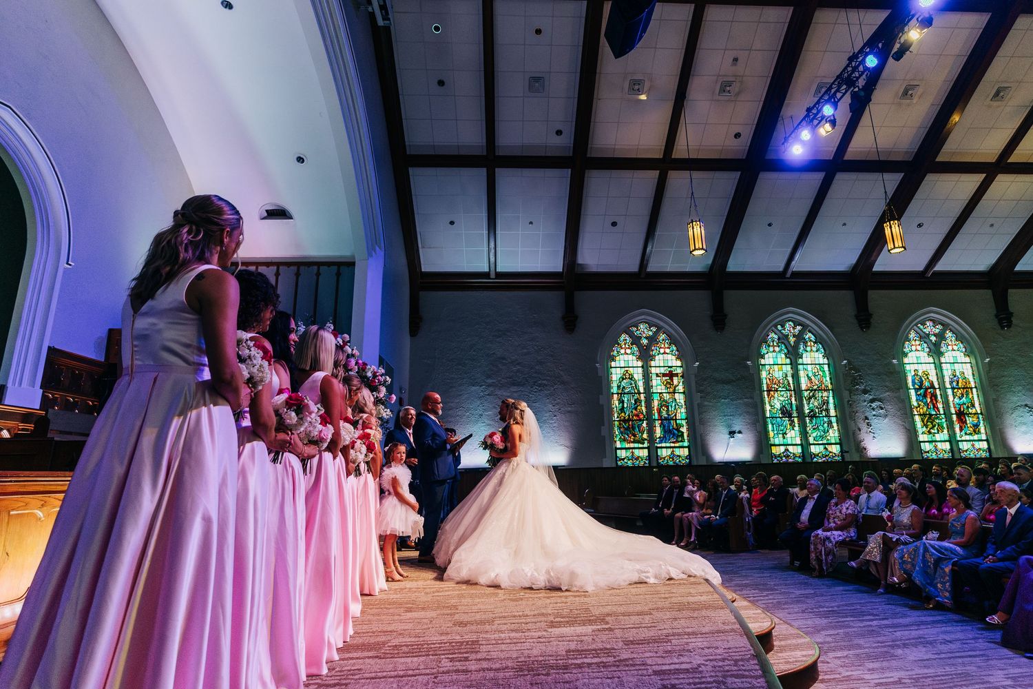 Bridesmaids in pink dresses watch as bride takes center stage during wedding ceremony in historic church with stained glass.