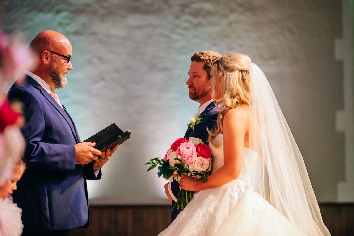 A wedding ceremony takes place as an officiant in a blue suit conducts the service with a bridal bouquet visible.