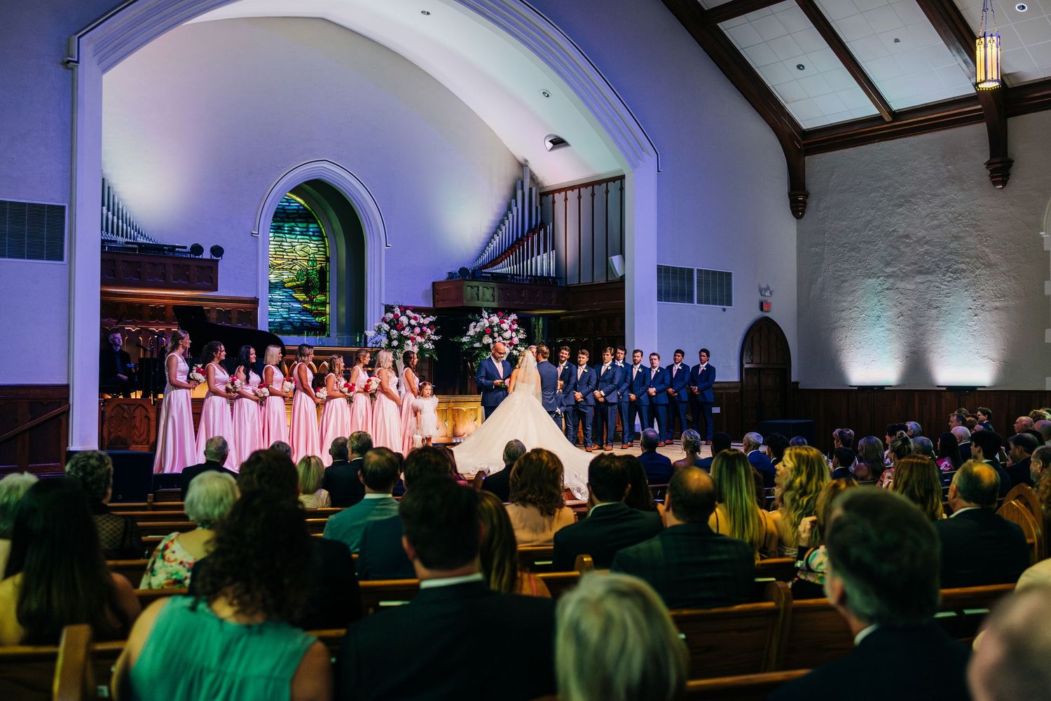 A grand church wedding ceremony with bridesmaids in pink dresses and groomsmen on stage in front of seated guests.