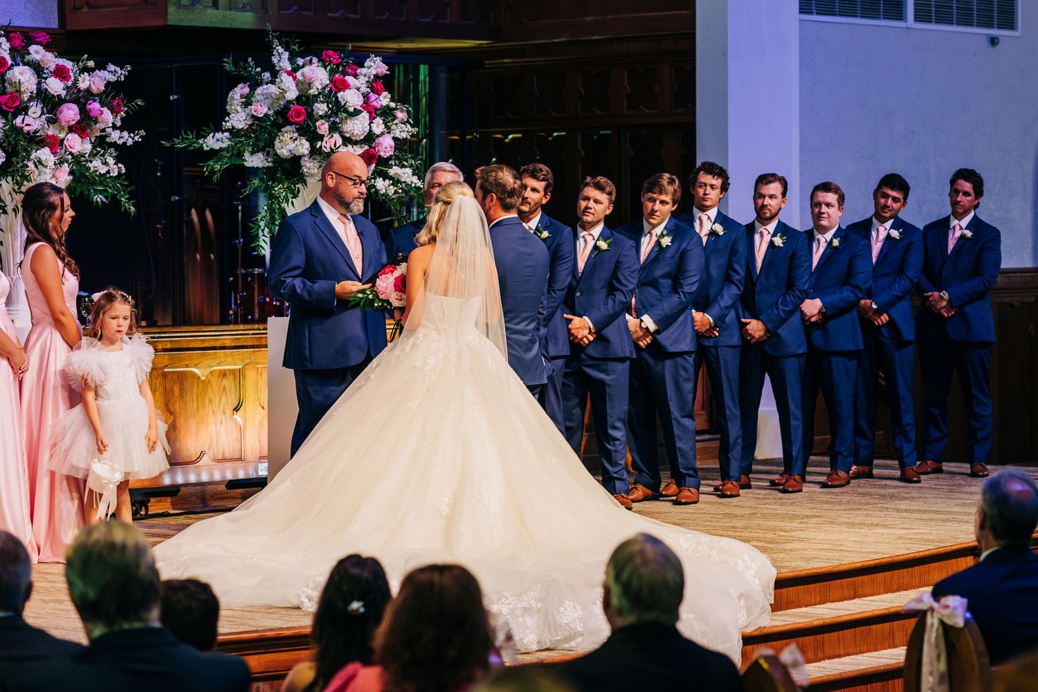 Wedding party in navy suits and pink dresses gather around couple during elegant church ceremony.