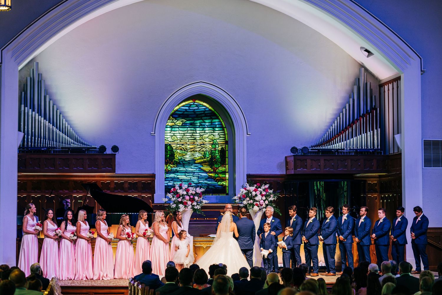 Wedding party stands in formation inside a church chapel with glowing purple lighting and stained glass window at the altar.