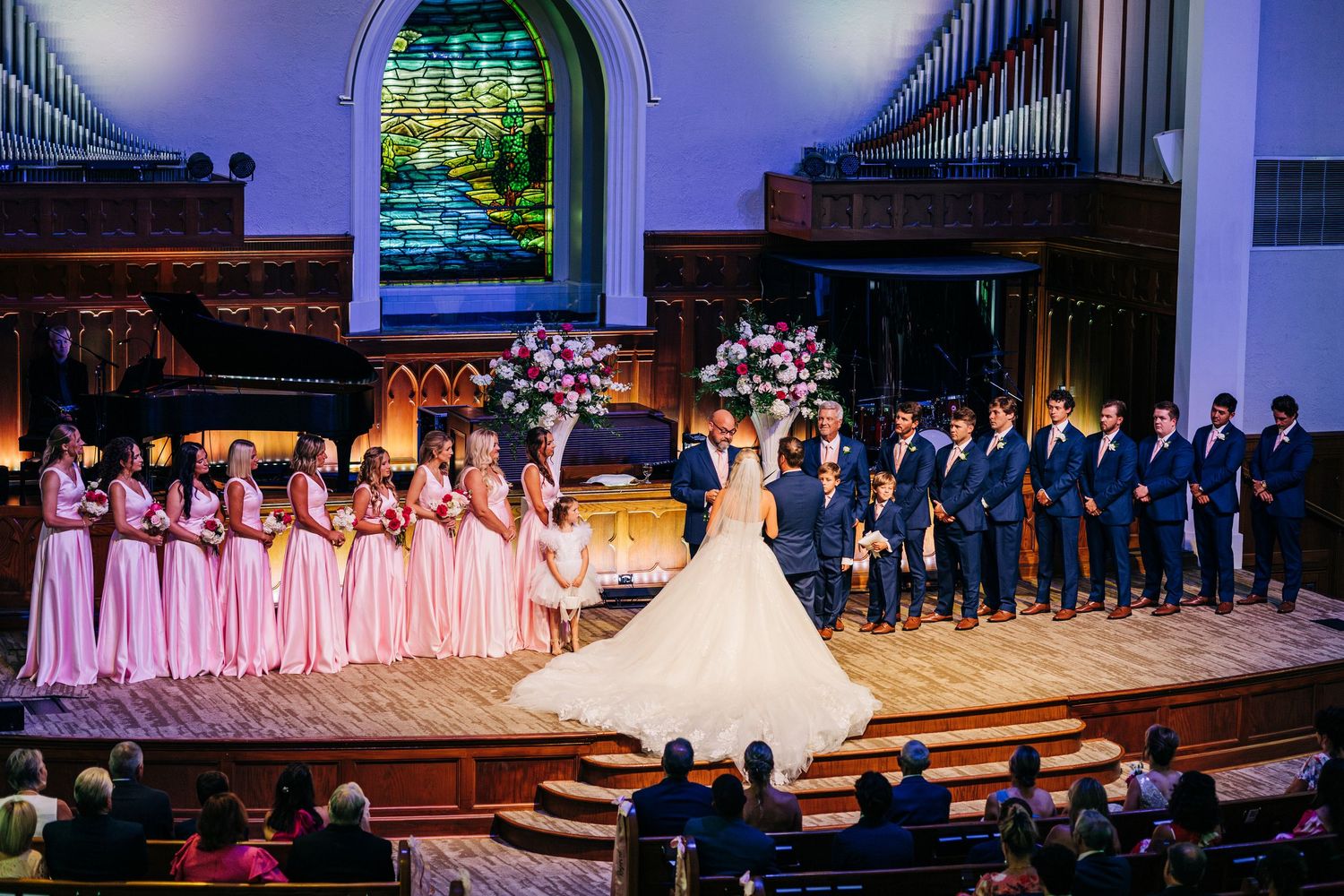 Wedding ceremony taking place in a grand church with bridesmaids in pink dresses and groomsmen in navy suits on stage.