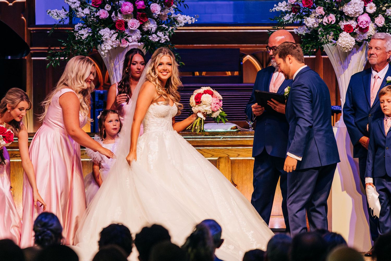 Wedding party in pink dresses and navy suits gather for a formal photograph at an elegant reception venue.
