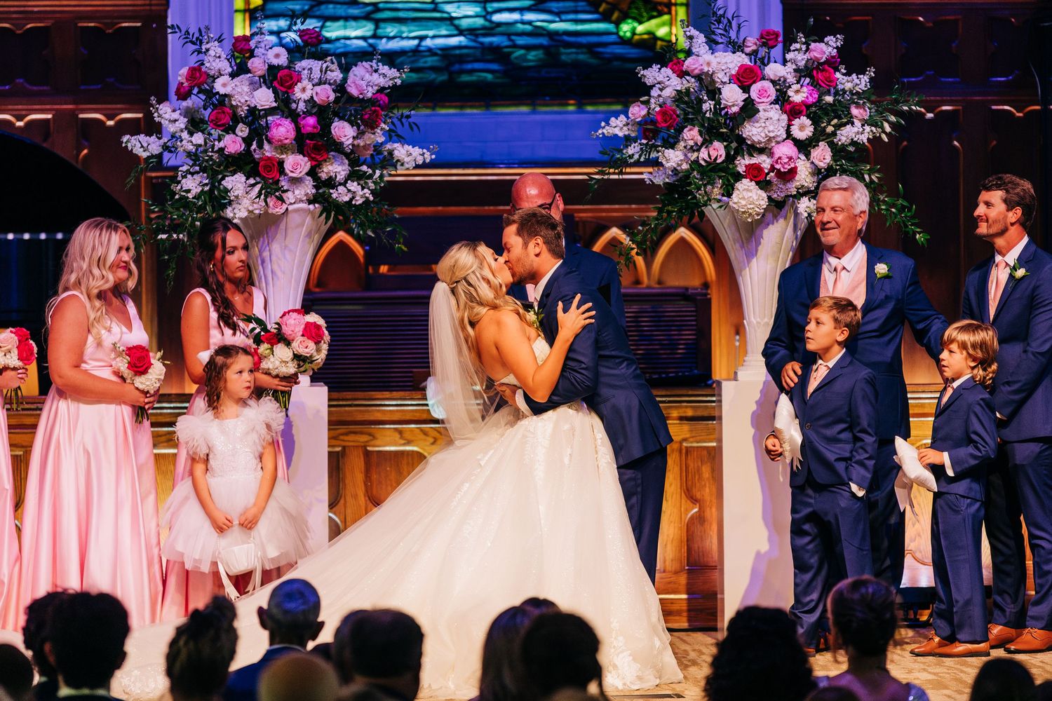 Wedding guests dance and celebrate during the reception under pink and purple mood lighting in a rustic venue.