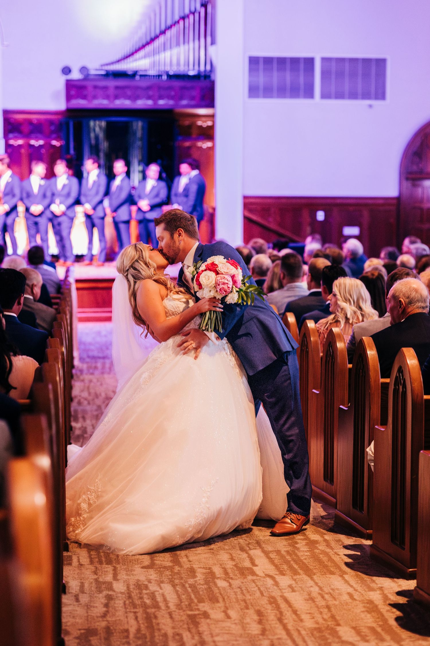 A couple shares a romantic kiss in a church aisle during their wedding ceremony with pink and purple lighting.