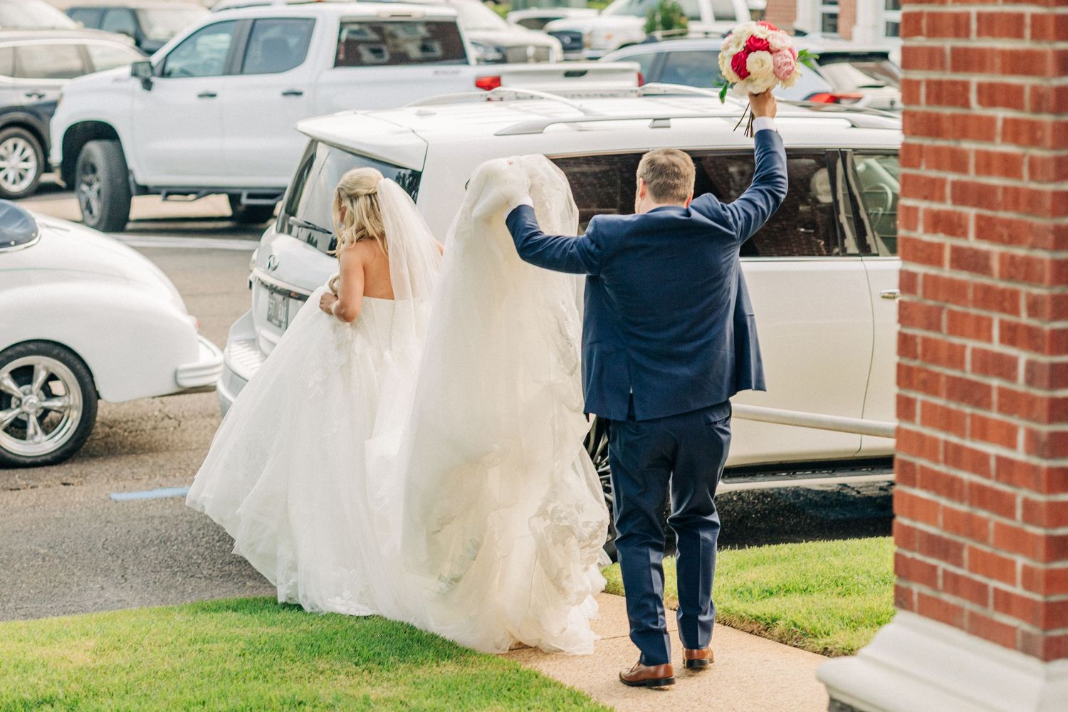 A person in a flowing white wedding dress walks in a parking lot next to red brick buildings.