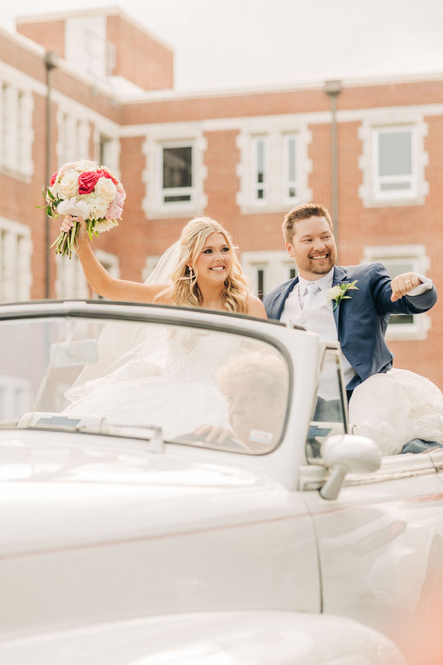 A joyful couple celebrates in a classic vintage car with a pink and white floral bouquet.