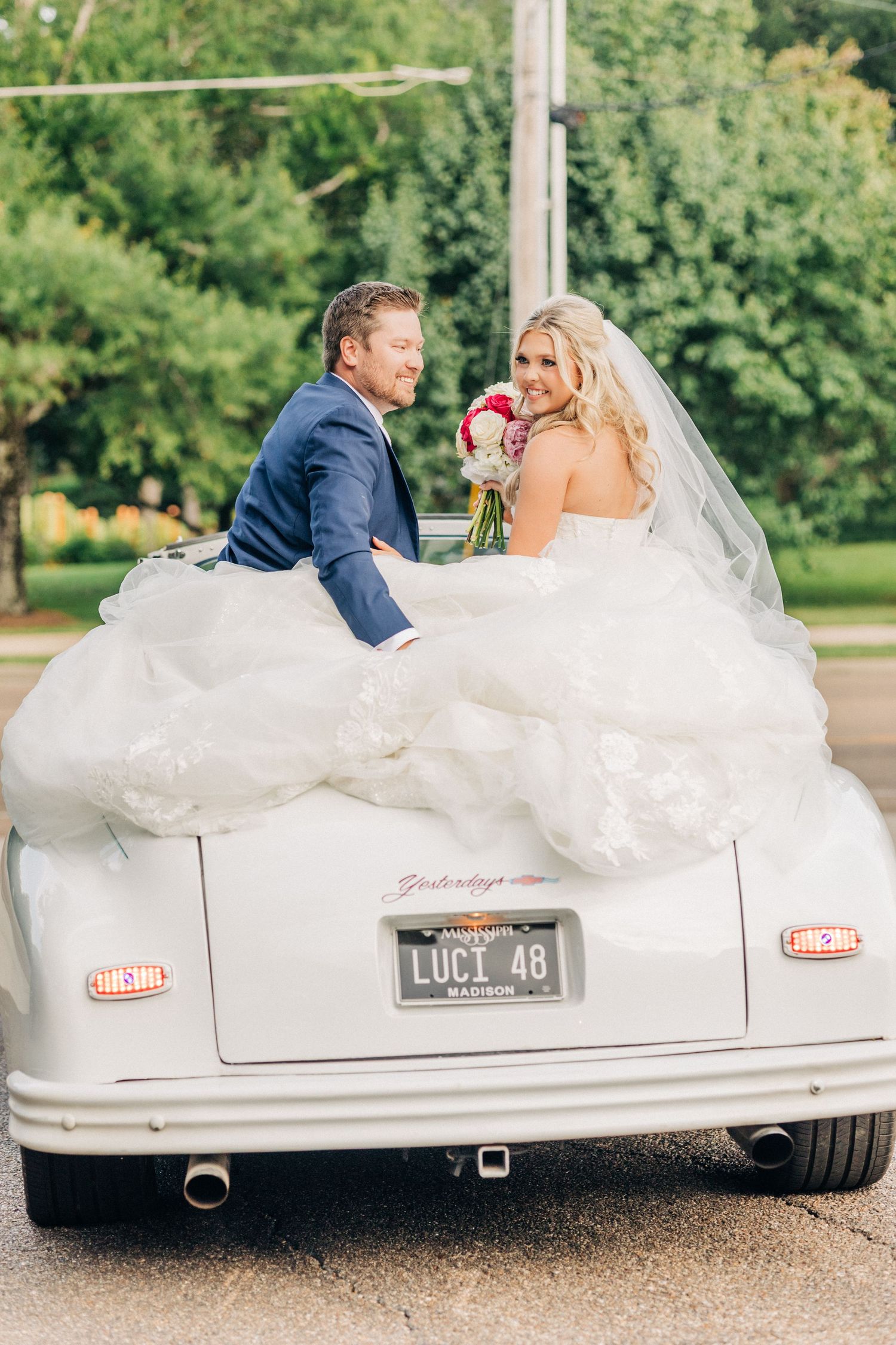A classic white convertible sports car parked on a gravel path beside greenery, decorated for a wedding celebration.