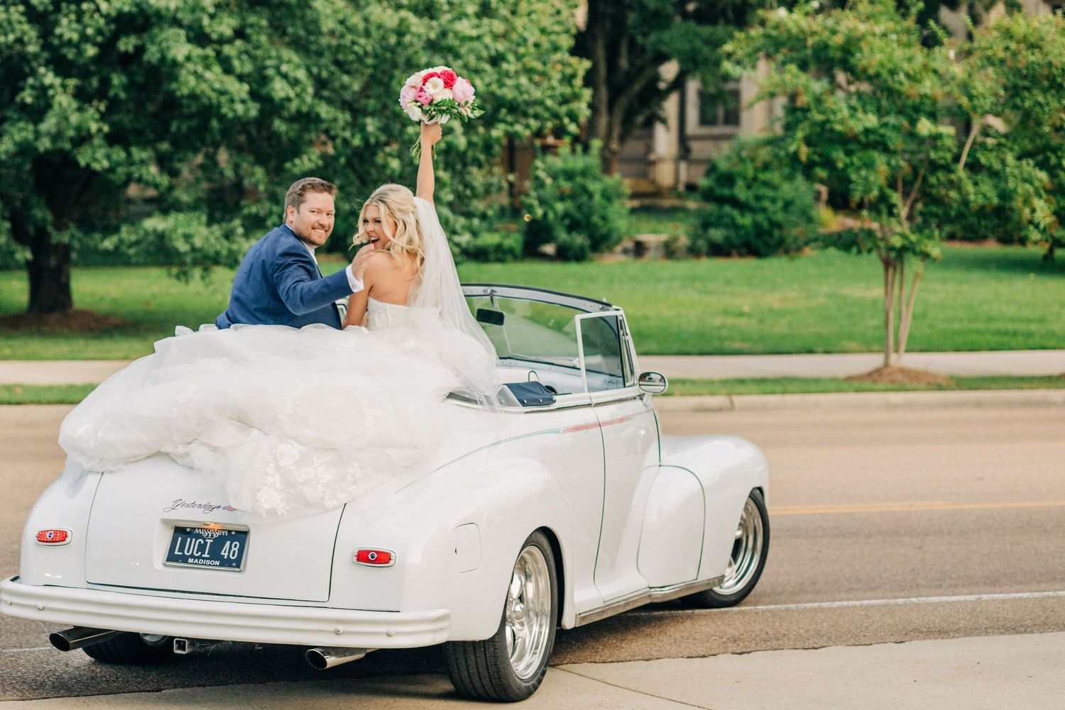 A white classic convertible car decorated as a wedding vehicle drives along a sunny road with lush greenery in the background.